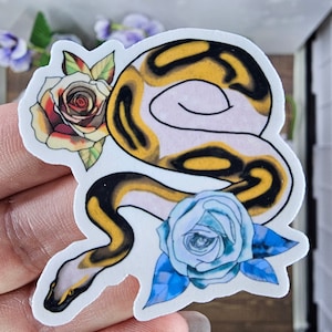 May include: A sticker featuring a yellow and black snake with a white belly, curled around a red rose and a blue rose. The snake is a ball python.