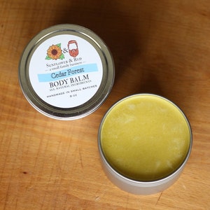 Könnte beinhalten: Eine silberne Dose mit einem weißen Etikett, auf dem "Sunflower & Red - a small family business - Cedar Forest Body Balm - All Natural Ingredients - Handmade in Small Batches - 8 oz" steht. Die Dose ist geöffnet und der Balsam ist gelb.