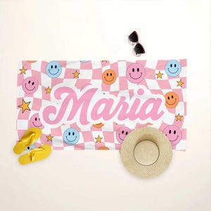 K&ouml;nnte beinhalten: Ein Strandtuch mit rosa-wei&szlig;em Karomuster, Smiley-Gesichtern und Sternen. Der Name "Maria" ist in rosa Schrift gedruckt. Gelbe Flip-Flops, ein Strohhut und eine Sonnenbrille sind ebenfalls zu sehen.