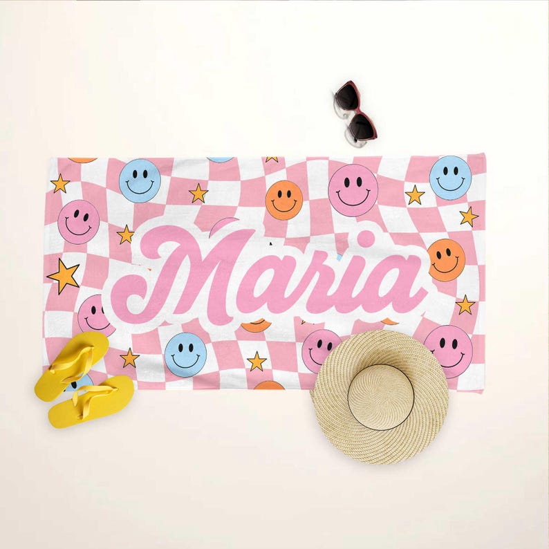 K&ouml;nnte beinhalten: Ein Strandtuch mit rosa-wei&szlig;em Karomuster, Smiley-Gesichtern und Sternen. Der Name "Maria" ist in rosa Schrift gedruckt. Gelbe Flip-Flops, ein Strohhut und eine Sonnenbrille sind ebenfalls zu sehen.