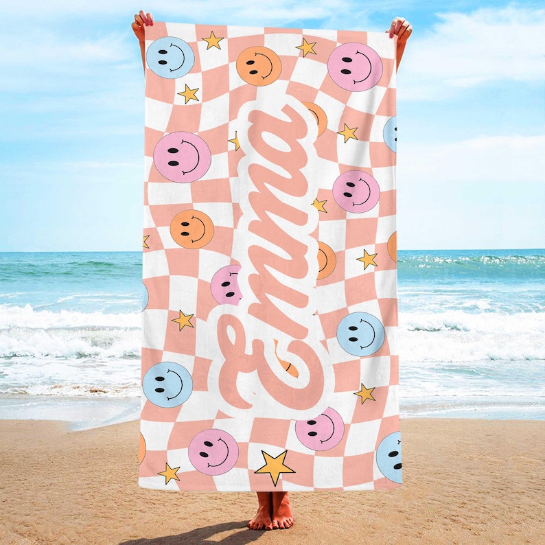 K&ouml;nnte beinhalten: Ein Strandtuch mit rosa-wei&szlig;em Karomuster, das Smiley-Gesichter in verschiedenen Farben und goldene Sterne zeigt. Der Name "Emma" ist vertikal in einer Schreibschrift gedruckt. Das Handtuch wird an einem Strand gehalten.
