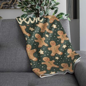 May include: A cozy throw blanket featuring a repeating pattern of gingerbread men and stars on a dark green background. The blanket is draped over a gray sofa, showcasing its festive design. The gingerbread men have smiling faces.
