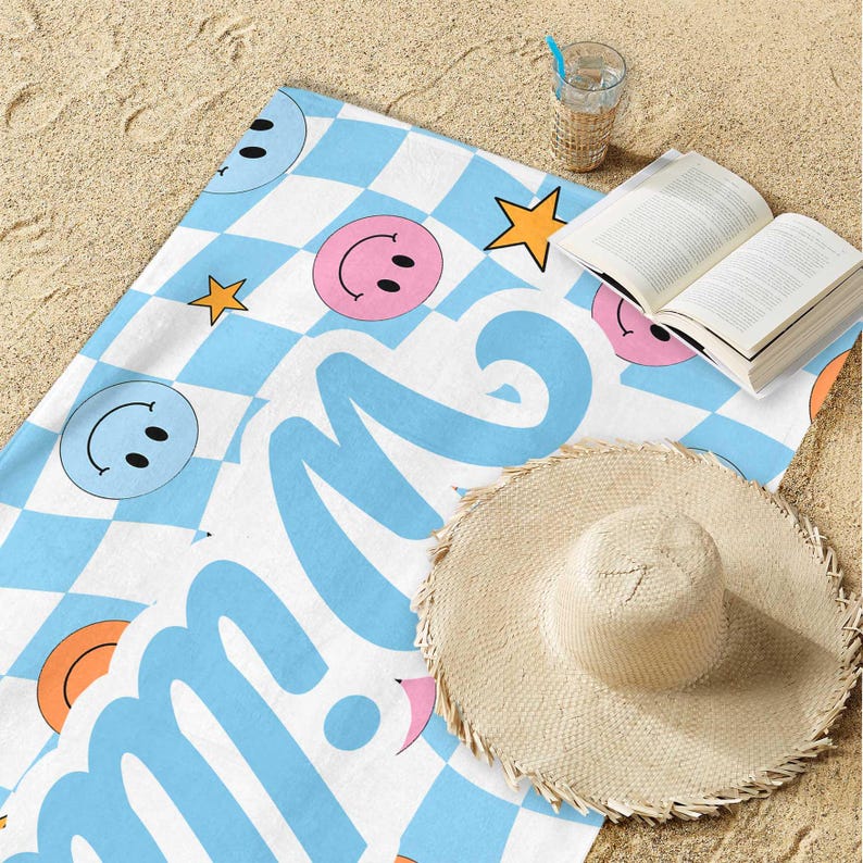 K&ouml;nnte beinhalten: Ein Strandtuch mit blau-wei&szlig;em Karomuster, Smiley-Gesichtern und Sternen. Das Wort "smile" ist in gro&szlig;en, hellblauen Buchstaben geschrieben. Ein Strohhut, ein offenes Buch und ein Glas Wasser mit Strohhalm liegen auf dem Sand.