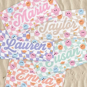K&ouml;nnte beinhalten: Mehrere personalisierte Strandt&uuml;cher mit Namen wie "Maria" und "Taylor" werden gezeigt. Jedes Handtuch hat ein Schachbrettmuster mit Smiley-Gesichtern und Sternen in verschiedenen Pastellfarben, darunter Rosa, Blau und Gr&uuml;n.