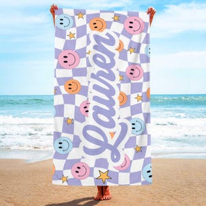K&ouml;nnte beinhalten: Strandtuch mit einem wei&szlig;-lila Schachbrettmuster, verziert mit bunten Smiley-Gesichtern und goldenen Sternen. Der Name "Lauren" ist vertikal in einer lila Schriftart aufgedruckt.