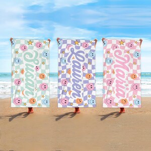 K&ouml;nnte beinhalten: Drei Strandt&uuml;cher mit Schachbrettmustern und Smiley-Gesichtern. Jedes Handtuch hat ein anderes Farbschema: Mintgr&uuml;n, Lavendel und Rosa. Die Namen "Susan", "Lauren" und "Maria" sind vertikal aufgedruckt, zusammen mit Sternen.