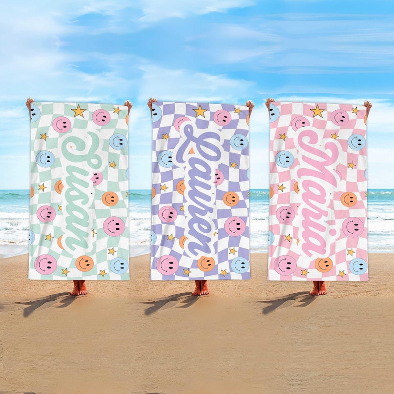 K&ouml;nnte beinhalten: Drei Strandt&uuml;cher mit Schachbrettmustern und Smiley-Gesichtern. Jedes Handtuch hat ein anderes Farbschema: Mintgr&uuml;n, Lavendel und Rosa. Die Namen "Susan", "Lauren" und "Maria" sind vertikal aufgedruckt, zusammen mit Sternen.