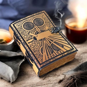 May include: A tan leather journal with a detailed black art deco design on the cover. The journal has a metal clasp and aged, yellowed pages. A small amount of smoke rises from the journal, suggesting it is warm.