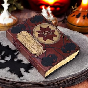 May include: A brown leather-bound journal with a gold embossed design featuring an Egyptian eye symbol and hieroglyphics. The journal is closed and resting on a gray stone surface.