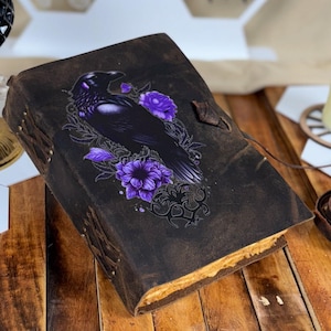 Może przedstawiać: Ciemnobrązowy skórzany dziennik z motywem kruka i fioletowych kwiatów. Dziennik ma postarzany wygląd, z widocznymi szwami i skórzanym zapięciem na pasek. Kruk jest przedstawiony ze szczegółami, otoczony fioletowymi kwiatami.