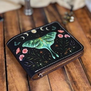 May include: Black wallet featuring a large green moth design with pink flowers, moons, and stars. The wallet has a brown leather interior and a white stitched border. The wallet is on a wooden surface.
