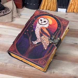 以下が含まれることがあります： 枝に止まっているフクロウのカラフルなイラストが描かれた革製のジャーナル。フクロウは白と茶色の羽根で、くちばしが黄色い。背景は濃い紫で、遠くには夕焼けが見える。ジャーナルには真鍮製の留め金が付いている。