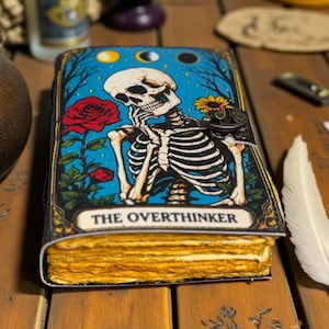May include: A journal with a cover featuring a skeleton in thought, a red rose, and the text "THE OVERTHINKER" against a blue background. The journal has aged, yellowed pages and a metal clasp.
