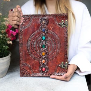 Puede incluir: Un diario de cuero marrón con un diseño en relieve decorativo y una fila de piedras preciosas de colores. El diario tiene un cierre de broche, mostrando los intrincados detalles.