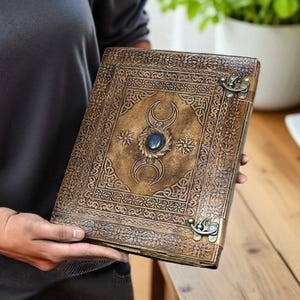 Pode incluir: Um di&aacute;rio de couro castanho com padr&otilde;es em relevo intrincados e uma gema azul central. O di&aacute;rio apresenta um fecho de metal e detalhes decorativos, sugerindo um estilo vintage ou artesanal. O design da capa inclui motivos de lua crescente e estrelas.