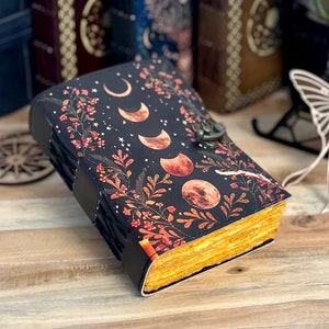 K&ouml;nnte beinhalten: Ein in schwarzes Leder gebundenes Tagebuch mit einer Grafik der Mondphasen und botanischen Illustrationen in Orange- und Rott&ouml;nen. Das Buch verf&uuml;gt &uuml;ber einen Metallverschluss und dicke, strukturierte Papierkanten in einem gelblichen Farbton.