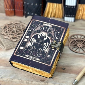 Könnte beinhalten: Ein schwarzes Lederjournal mit einer goldenen und schwarzen Illustration der Teufels-Tarotkarte. Das Journal hat einen Verschluss mit einer Schließe und einen vergilbten Buchschnitt.