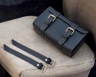 May include: A black leather tool roll with two adjustable straps and buckles. The roll is sitting on a light brown fabric surface.