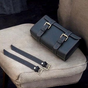 May include: A black leather tool roll with two adjustable straps and buckles. The roll is sitting on a light brown fabric surface.
