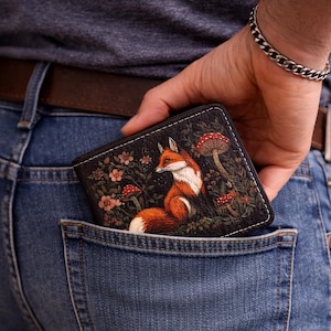 Könnte beinhalten: Schwarzes Portemonnaie mit einer detaillierten Illustration eines Fuchses in einer Waldszene mit Pilzen und Blumen. Das Portemonnaie wird in die Gesäßtasche einer Jeans gesteckt. Der Fuchs ist orange, weiß und braun.
