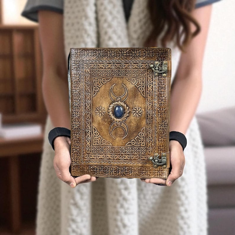 Pode incluir: Um di&aacute;rio de couro marrom com padr&otilde;es em relevo intrincados e um design central com uma pedra preciosa azul e luas crescentes. O di&aacute;rio tem um fecho de metal, mostrando o artesanato detalhado.