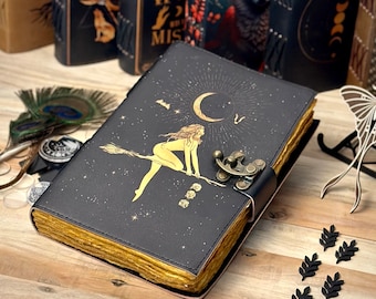 Puede incluir: Un diario de cuero negro con un cierre dorado y un diseño de una mujer montando una escoba bajo una luna creciente. El diario tiene una encuadernación dorada y está decorado con estrellas doradas.