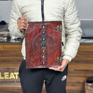 May include: A brown leather journal with a decorative embossed design and a row of colorful gemstones down the center. The journal has a metal clasp and is held by a person's hands.