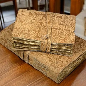 May include: Two antique-style leather-bound journals with gold-leaf pages and intricate floral patterns. The journals are closed and stacked on top of each other.