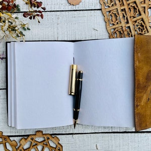 May include: A brown leather bound journal with blank white pages, two pens, one gold and one black, are laying open on a white wooden surface.