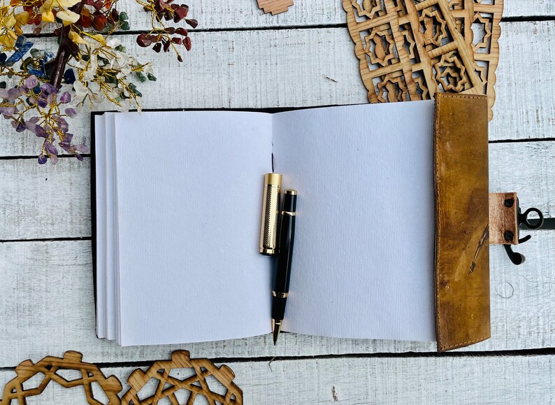 May include: A brown leather bound journal with blank white pages, two pens, one gold and one black, are laying open on a white wooden surface.