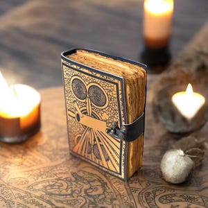 May include: A vintage journal featuring an owl design on the cover. The cover is gold with black detailing and a black leather strap. The pages have a worn appearance. Candles and a map are visible in the background, creating a mystical atmosphere.