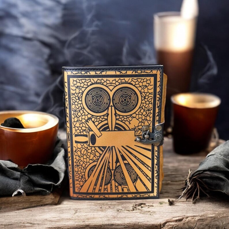 May include: A tan leather journal with a black border and a decorative design featuring a stylized face. The journal has a metal clasp and is displayed on a wooden surface. The design includes circular and linear elements.
