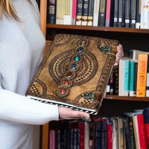 Puede incluir: Un diario de cuero marr&oacute;n con intrincados dise&ntilde;os en relieve y una fila de gemas coloridas. El diario tiene un cierre de broche y un borde decorativo. La portada presenta un patr&oacute;n circular central con l&iacute;neas radiales y peque&ntilde;os detalles circulares.