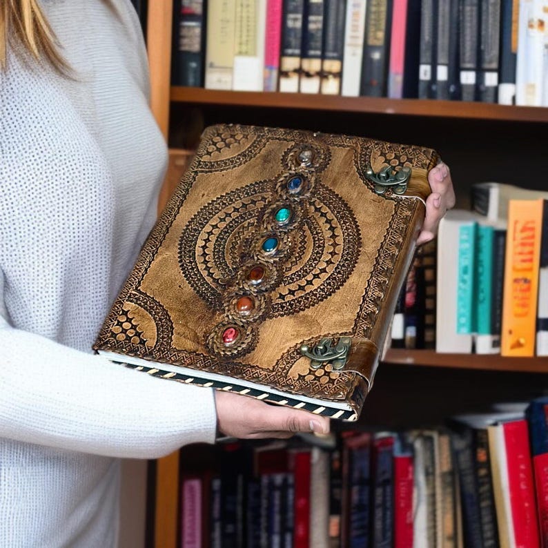 Puede incluir: Un diario de cuero marr&oacute;n con intrincados dise&ntilde;os en relieve y una fila de gemas coloridas. El diario tiene un cierre de broche y un borde decorativo. La portada presenta un patr&oacute;n circular central con l&iacute;neas radiales y peque&ntilde;os detalles circulares.