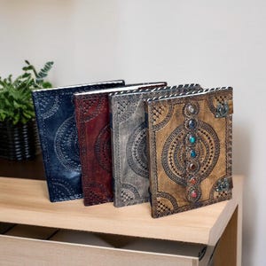 Puede incluir: Cuatro diarios de cuero en tonos azul, burdeos, gris y marr&oacute;n, cada uno con patrones circulares en relieve. El diario marr&oacute;n presenta una fila vertical de piedras preciosas de colores y un cierre de metal. Los diarios se exhiben sobre una superficie de madera.