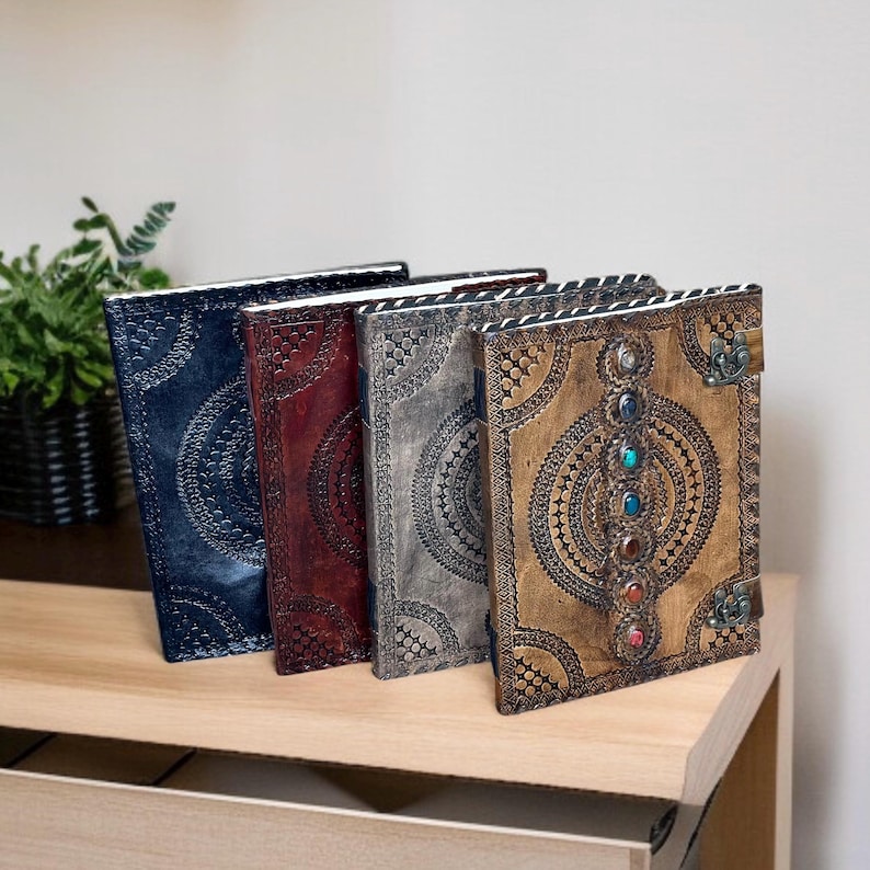 Puede incluir: Cuatro diarios de cuero en tonos azul, burdeos, gris y marr&oacute;n, cada uno con patrones circulares en relieve. El diario marr&oacute;n presenta una fila vertical de piedras preciosas de colores y un cierre de metal. Los diarios se exhiben sobre una superficie de madera.