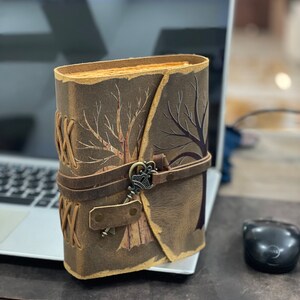 May include: A brown leather journal with a tree design and a keyhole closure. The journal has a brown leather strap and a keyhole with a metal key.