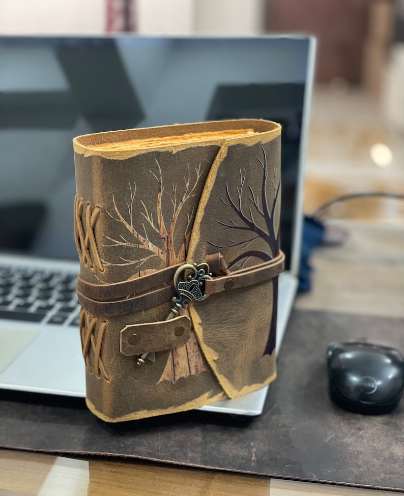 May include: A brown leather journal with a tree design and a keyhole closure. The journal has a brown leather strap and a keyhole with a metal key.
