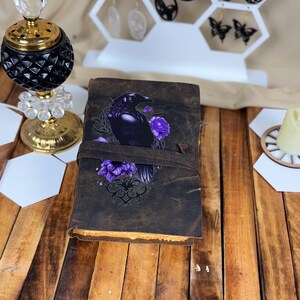 以下が含まれることがあります： カラスと紫の花のデザインが施された革張りのジャーナル。ジャーナルは、ストラップ留めの濃い茶色の表紙です。カラスは黒で紫色のアクセントがあり、花は鮮やかな紫色です。ページの端は黄色です。