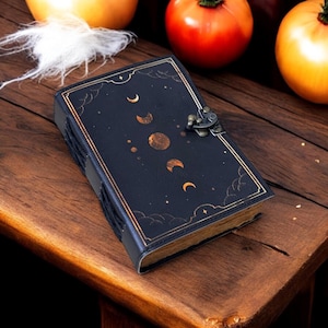 K&ouml;nnte beinhalten: Ein schwarzes Lederjournal mit einem goldenen Mond- und Sternenmuster. Das Journal hat einen Verschluss mit einer Schlie&szlig;e und liegt auf einer Holzoberfl&auml;che.