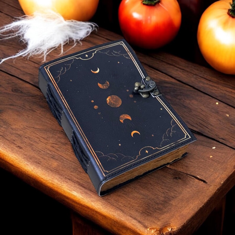 K&ouml;nnte beinhalten: Ein schwarzes Lederjournal mit einem goldenen Mond- und Sternenmuster. Das Journal hat einen Verschluss mit einer Schlie&szlig;e und liegt auf einer Holzoberfl&auml;che.