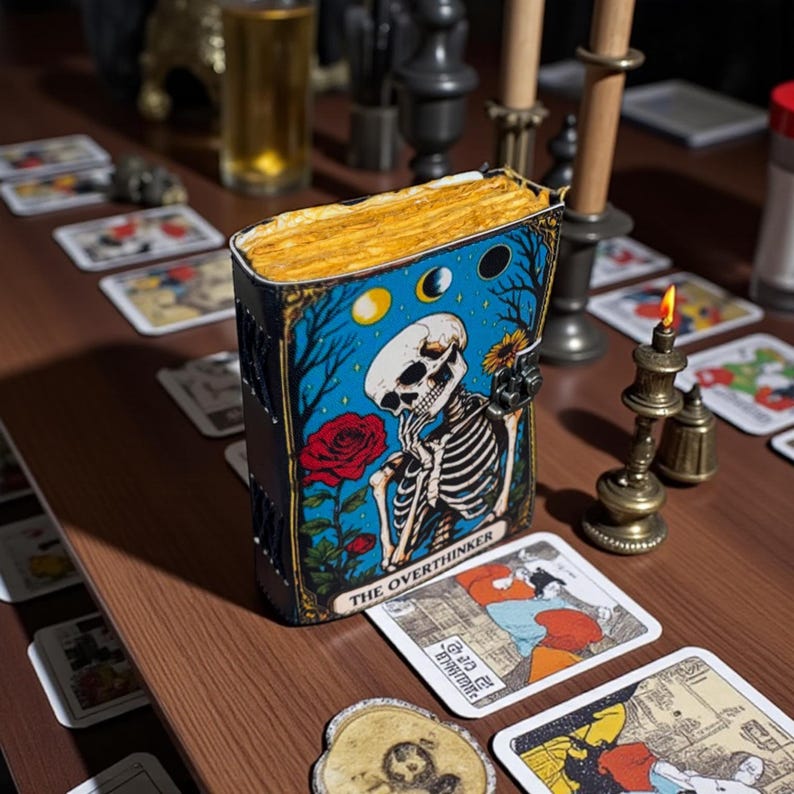 May include: A decorative tarot card box with a blue background featuring a skeleton, a red rose, and the text "THE OVERTHINKER." The box is surrounded by tarot cards and a lit candle. The box has a gold-colored top.