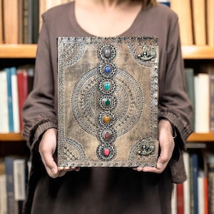 Puede incluir: Un diario de cuero marr&oacute;n con un marco met&aacute;lico decorativo y una serie de piedras preciosas de colores. El diario presenta un dise&ntilde;o en relieve y un cierre met&aacute;lico. Las piedras preciosas est&aacute;n dispuestas verticalmente.