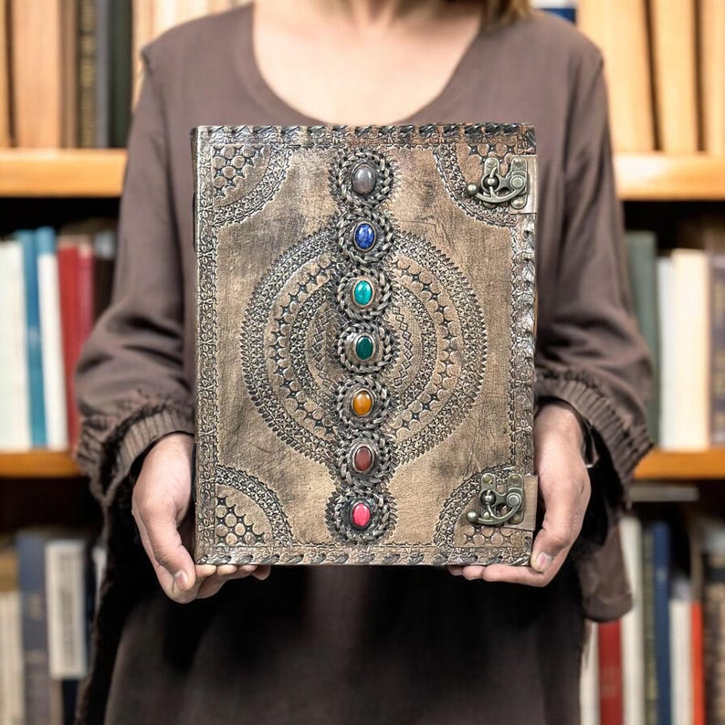 Puede incluir: Un diario de cuero marr&oacute;n con un marco met&aacute;lico decorativo y una serie de piedras preciosas de colores. El diario presenta un dise&ntilde;o en relieve y un cierre met&aacute;lico. Las piedras preciosas est&aacute;n dispuestas verticalmente.