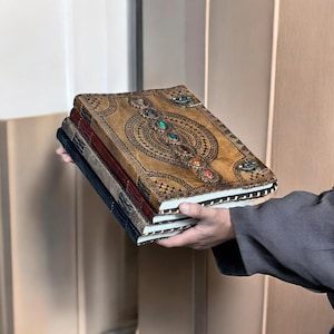 Puede incluir: Una pila de tres diarios encuadernados en cuero. El diario superior es de color canela con un dise&ntilde;o en relieve ornamentado y detalles de piedras preciosas de colores. Los otros dos diarios son de color azul oscuro y burdeos.