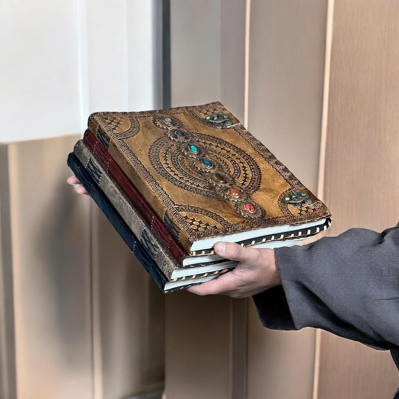 Puede incluir: Una pila de tres diarios encuadernados en cuero. El diario superior es de color canela con un dise&ntilde;o en relieve ornamentado y detalles de piedras preciosas de colores. Los otros dos diarios son de color azul oscuro y burdeos.