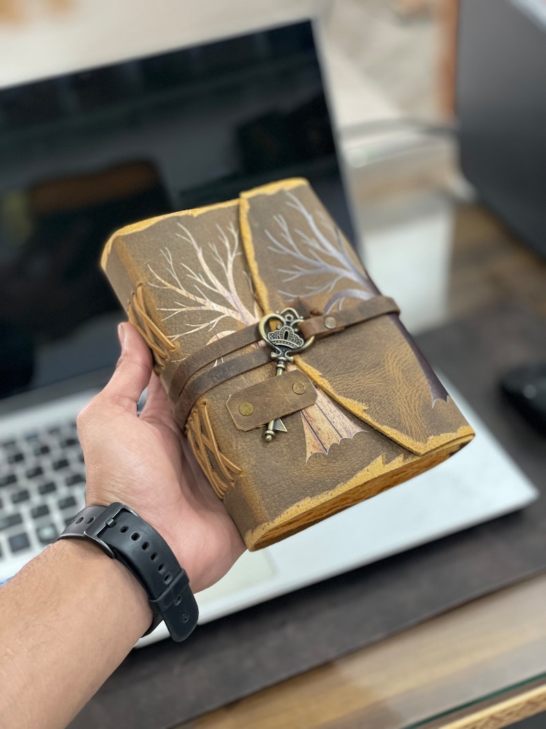 May include: A brown leather journal with a tree design and a metal key charm. The journal has a strap closure and is held in a hand.