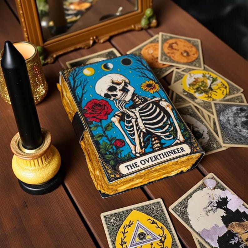 May include: A tarot deck with a cover featuring a skeleton, roses, and the text "THE OVERTHINKER." The deck is surrounded by other tarot cards, a black candle, and a gold-colored candle holder. The background includes a small mirror.