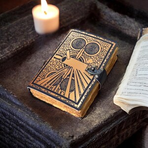 May include: A decorative journal with a gold and black Art Deco design on the cover. The journal has a black leather strap with a metal clasp. The pages are aged, and a lit candle is in the background.