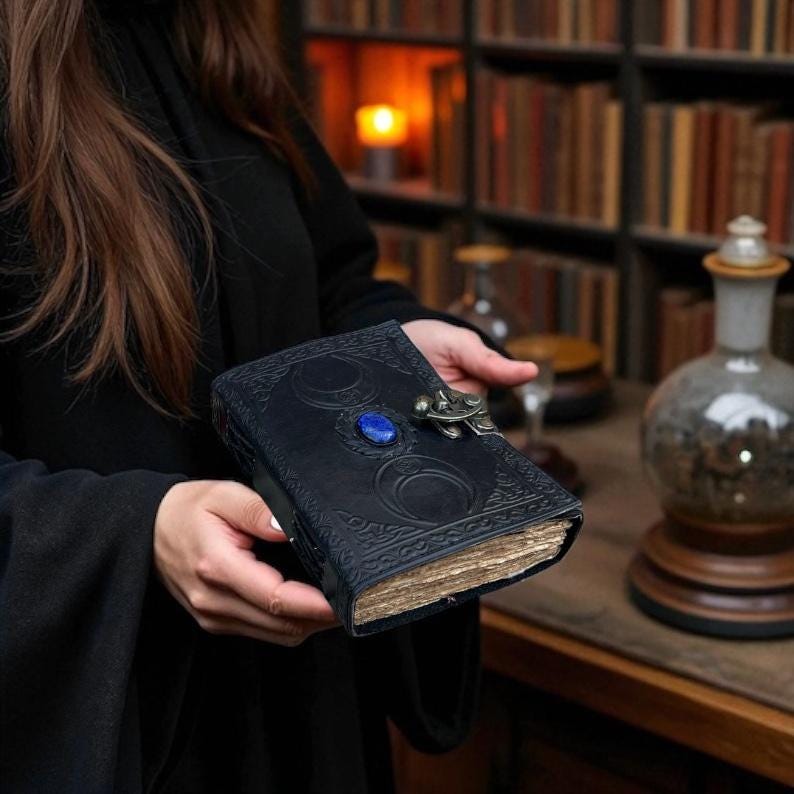 Pode incluir: Um di&aacute;rio em couro azul escuro com desenhos em relevo de lua crescente e estrelas. O di&aacute;rio apresenta uma pedra preciosa azul e um fecho decorativo. As p&aacute;ginas envelhecidas sugerem um estilo vintage.